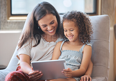 Happy little girl and loving mother laughing while sitting together on couch and using digital tablet to watch a movie or do a video call with family. Parent sitting with child while enjoying some online entertainment