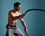 Handsome young hispanic man training with battle ropes in studio isolated against a blue background. Mixed race shirtless male athlete exercising or working out to increase his strength and fitness