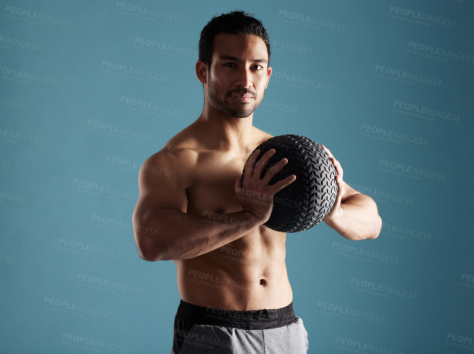 Buy stock photo Handsome young hispanic man training with a medicine ball in studio isolated against a blue background. Mixed race shirtless male athlete exercising or working out to increase his strength and fitness