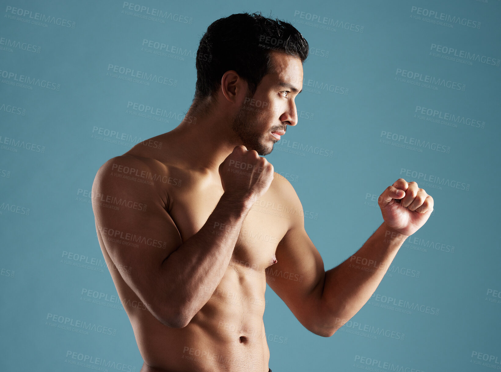 Buy stock photo Handsome young hispanic man standing shirtless in a boxer pose in studio isolated against a blue background. Mixed race topless male athlete, ready for boxing or a fist fight. A confident sportsman