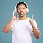 Handsome young mixed race man looking surprised and listening to music while standing in studio isolated against a blue background. Hispanic male remembering a song while using wireless headphones