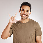 Handsome young mixed race man gesturing "rock on" while standing in studio isolated against a grey background. Hispanic male showing the sign language of "I love you" to show affection or romance