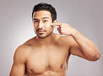 Handsome young mixed race man shirtless in studio isolated against a grey background. Hispanic male applying moisturizer lotion to his face. Looking confident and happy with his daily skincare regime