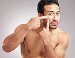 Handsome young mixed race man posing shirtless in studio isolated against a grey background. Hispanic male popping pimples or zits on his face. No outbreaks in the plans for his skincare regime