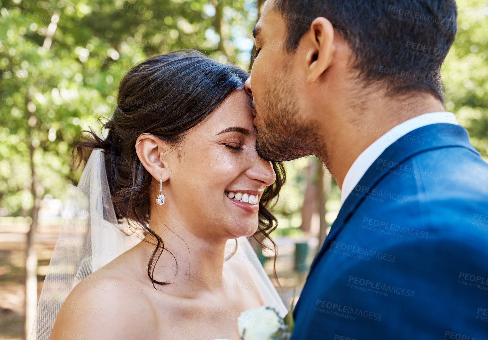 Buy stock photo Beautiful young couple on their wedding day. Groom kissing his joyful bride on the forehead while they stand together on a sunny day