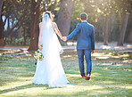 Rear view shot of newlywed couple walking together in nature. Bride and groom holding hands while walking together on summer day in nature. Wife following husband as he leads the way