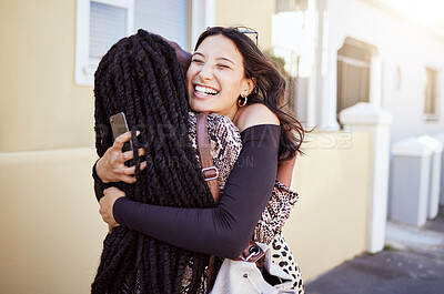 Buy stock photo Cheerful young woman holding her smartphone while hugging her friend on sidewalk. Female friends embracing and greeting each other on city street