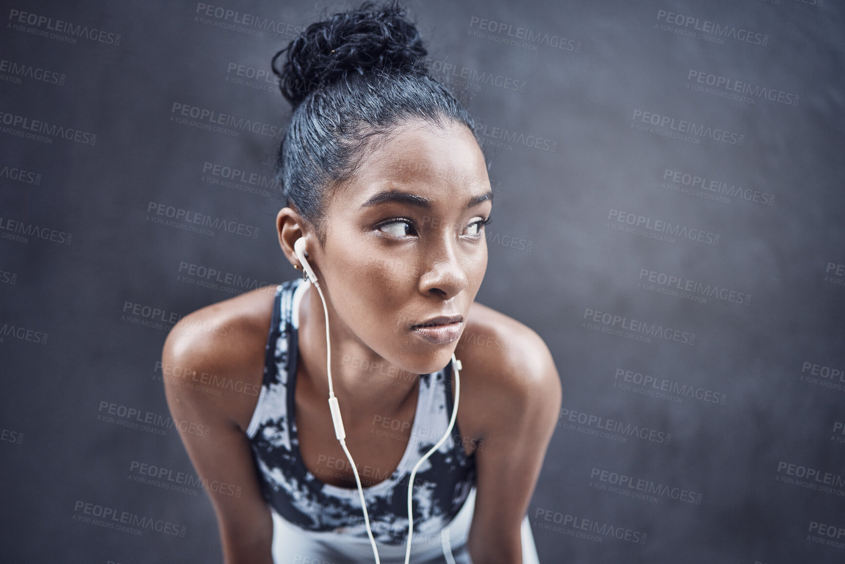Buy stock photo One active young mixed race woman wearing earphones and taking a rest break after run or jog exercise outdoors. Focused female athlete looking sweaty and tired but determined after challenging workout against a dark background
