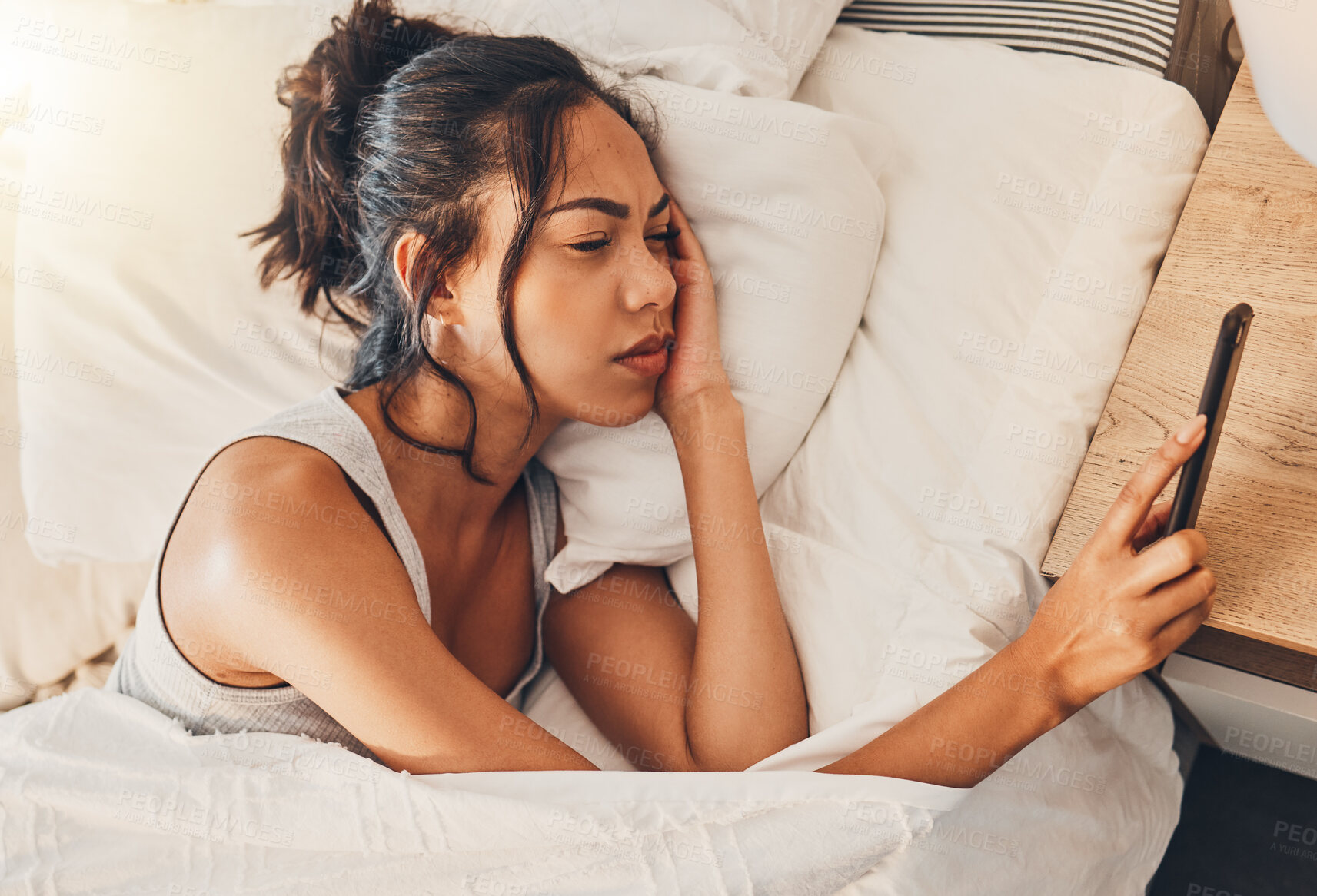 Buy stock photo A young mixed race woman looking depressed while scrolling social media and lying in bed. An attractive Hispanic female using her cellphone and looking exhausted while resting in her bedroom