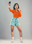 Full length mixed race woman taking selfies while isolated against grey studio background with copyspace. Young hispanic standing alone and pouting while taking pictures for social media on cellphone