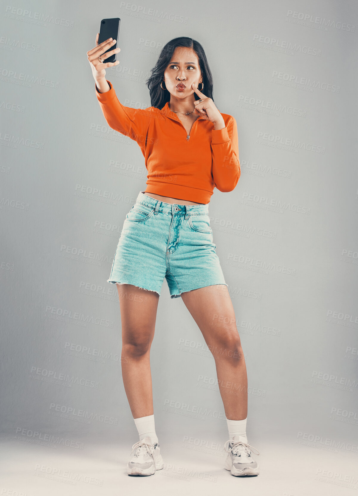 Buy stock photo Full length mixed race woman taking selfies while isolated against grey studio background with copyspace. Young hispanic standing alone and pouting while taking pictures for social media on cellphone