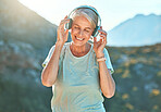 Active senior woman wearing headphones and listening to music with her eyes closed. Mature woman standing outdoors against mountain background
