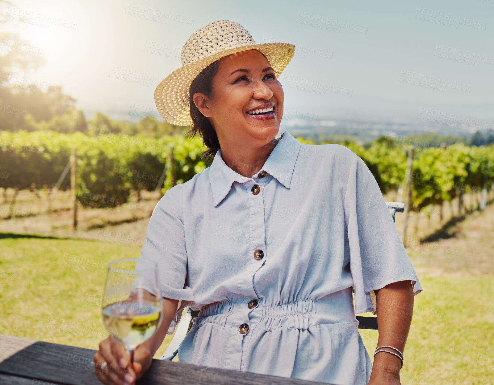 Buy stock photo One smiling mature mixed race woman enjoying a wine tasting day on a farm. Happy hispanic woman wearing a hat while sitting alone on a vineyard. Woman holding a glass of white wine during the weekend