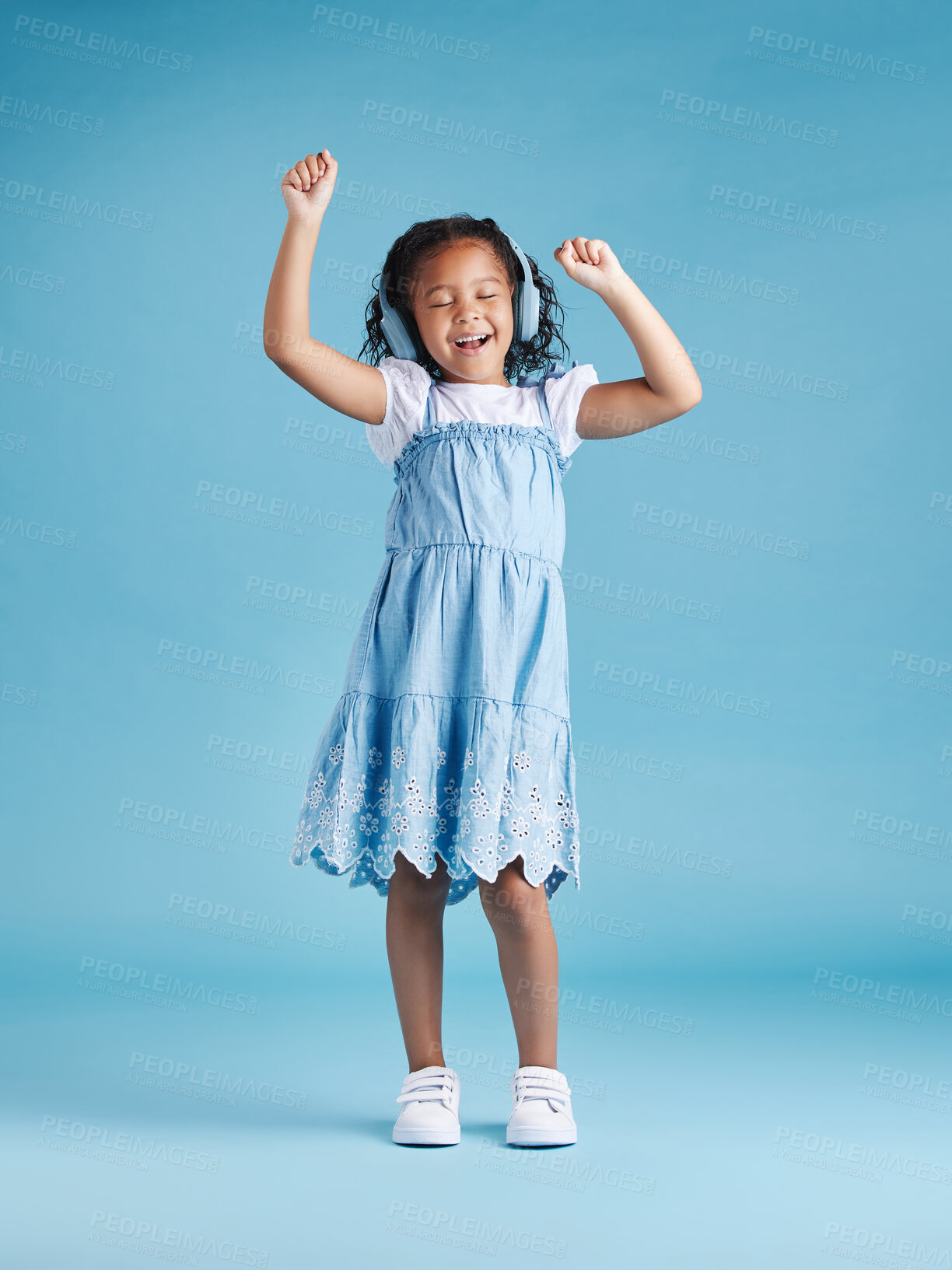 Buy stock photo Full length of an adorable little hispanic girl dancing with her hands up looking happy while listening to music with wireless headphones enjoying her favourite song against a blue studio background