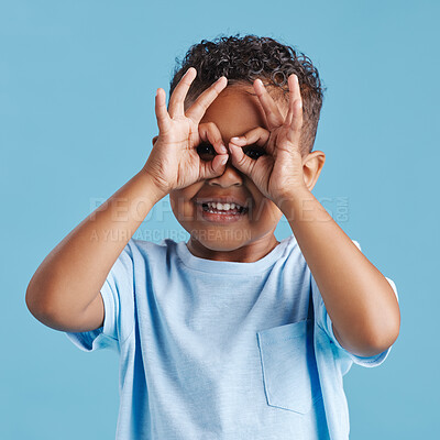 Portrait of inquisitive nosy little boy looking through fingers shaped like binoculars against a blue studio background. Curious child exploring