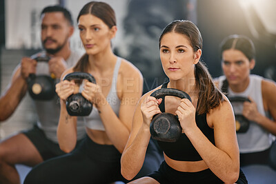 Active young caucasian woman doing kettlebell squat exercises while training together with a group of people in a gym. Focused athletes challenging themselves by holding heavy weights to build muscle and endurance during a workout in a fitness class