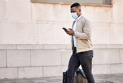 Buy stock photo Black businessman travelling alone.A african american businessman walking around town with his luggage while looking at his smartphone and wearing a mask to protect himself from the corona virus in the city