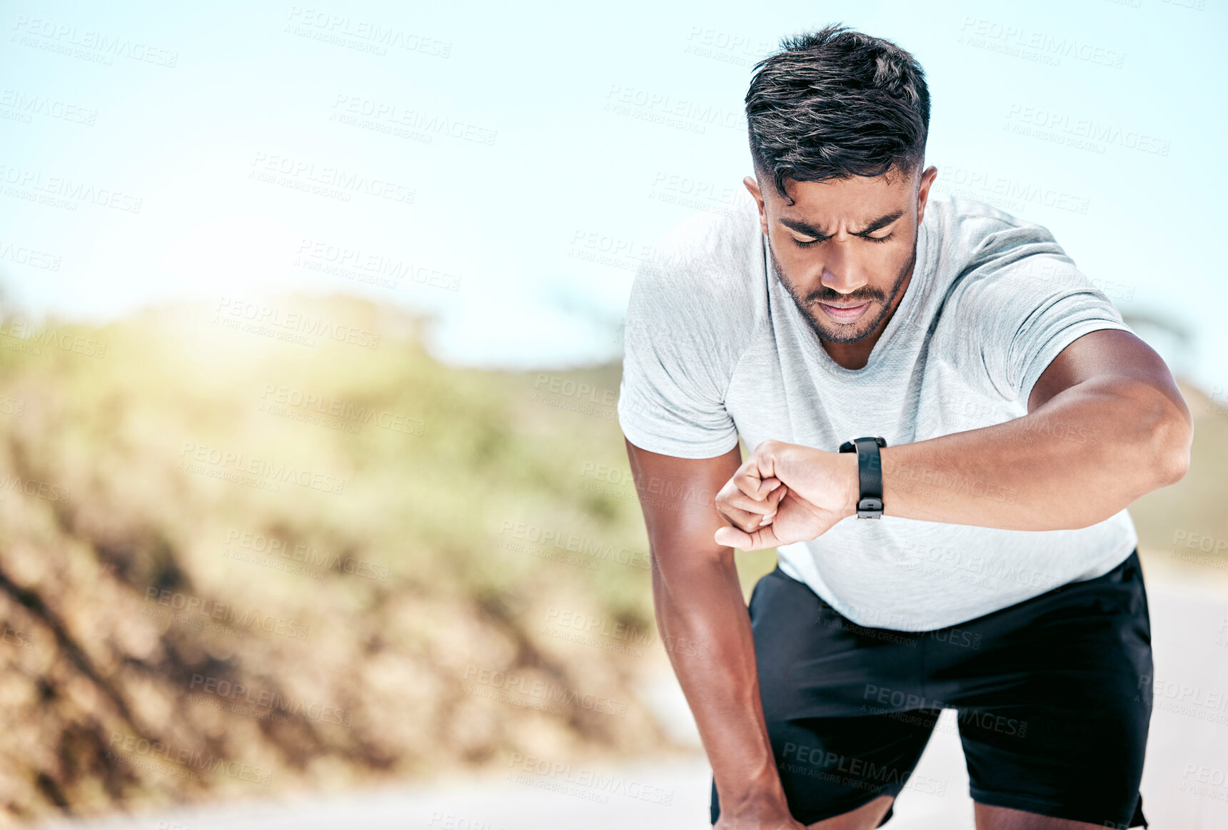 Buy stock photo Athletic young mixed race man looking at his watch while exercising outdoors. Handsome hispanic resting while checking the time on his smartwatch during a workout. Tracking progress while working out