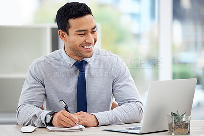 Young happy mixed race businessman writing in a diary while working on a laptop alone in an office at work. One content hispanic male businessperson taking notes in a book while sitting at a desk at work