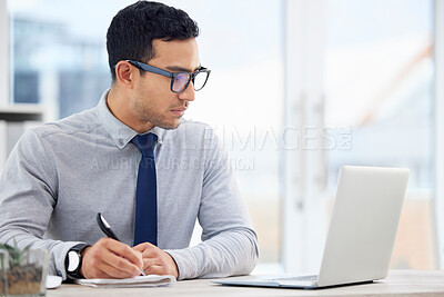 Focused mixed race businessman writing in a diary while working on a laptop alone in an office at work. One serious hispanic male businessperson making a list in a notebook while sitting at a desk at work