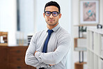 Young serious mixed race businessman standing with his arms crossed alone in an office at work. Headshot of a confident hispanic businessperson wearing glasses while standing at work