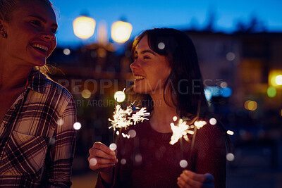 Young woman smiling at her friend and celebrating playing with fireworks. Two friends on holiday together celebrating with fireworks at night.Happy woman on holiday with friend playing with fireworks