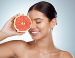 Closeup of smiling woman holding grapefruit over her eye while posing topless. Happy caucasian model isolated against a grey background in a studio with smooth skin, fresh healthy skincare routine
