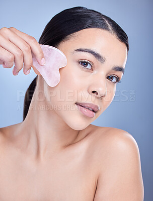 Buy stock photo Studio portrait of a beautiful mixed race woman using a rose quartz gua sha for deep penetration cell renewal. Young hispanic woman using anti ageing tool against blue copyspace background