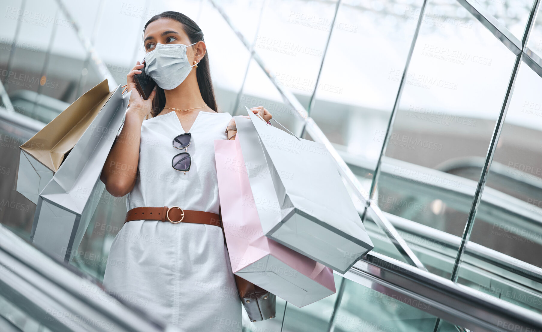 Buy stock photo One young mixed race woman wearing a medical face mask and talking on a cellphone while on an escalator after a shopping spree. Fashionable hispanic carrying retail bags after buying in a mall during Covid-19 pandemic