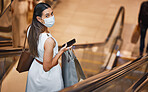 Portrait of a young mixed race woman wearing a medical face mask and holding a cellphone and shopping bags while on an escalator. Serious hispanic woman looking back over shoulder while carrying retail bags after buying in a mall during Covid-19 pandemic