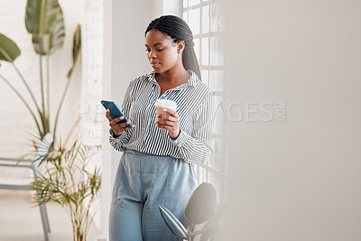 Buy stock photo African american businesswoman using her phone while drinking a coffee alone in an office at work. One black female businessperson using social media on her cellphone and holding a coffee cup on a break standing at work