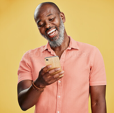 One happy African American man standing against a yellow studio background, holding and using his cellphone to browse the internet. Smiling black man laughing while browsing his phone for social media