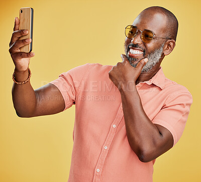 One happy trendy mature African American man taking selfies on his cellphone against a yellow studio background. Fashionable black man standing and posing while taking photographs for social media