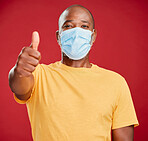 A african american making wearing mask to stop the spread of coronavirus and standing against a red studio background while showing a thumbs up gesture with his hand. Black male suggesting that everything will be alright. Look after our health wearing a m