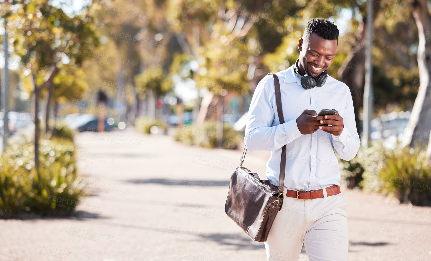 Buy stock photo Happy African American man wearing headphones and texting on a smartphone while out in the city on daily commute. Black male smiling while checking social media and walking outside