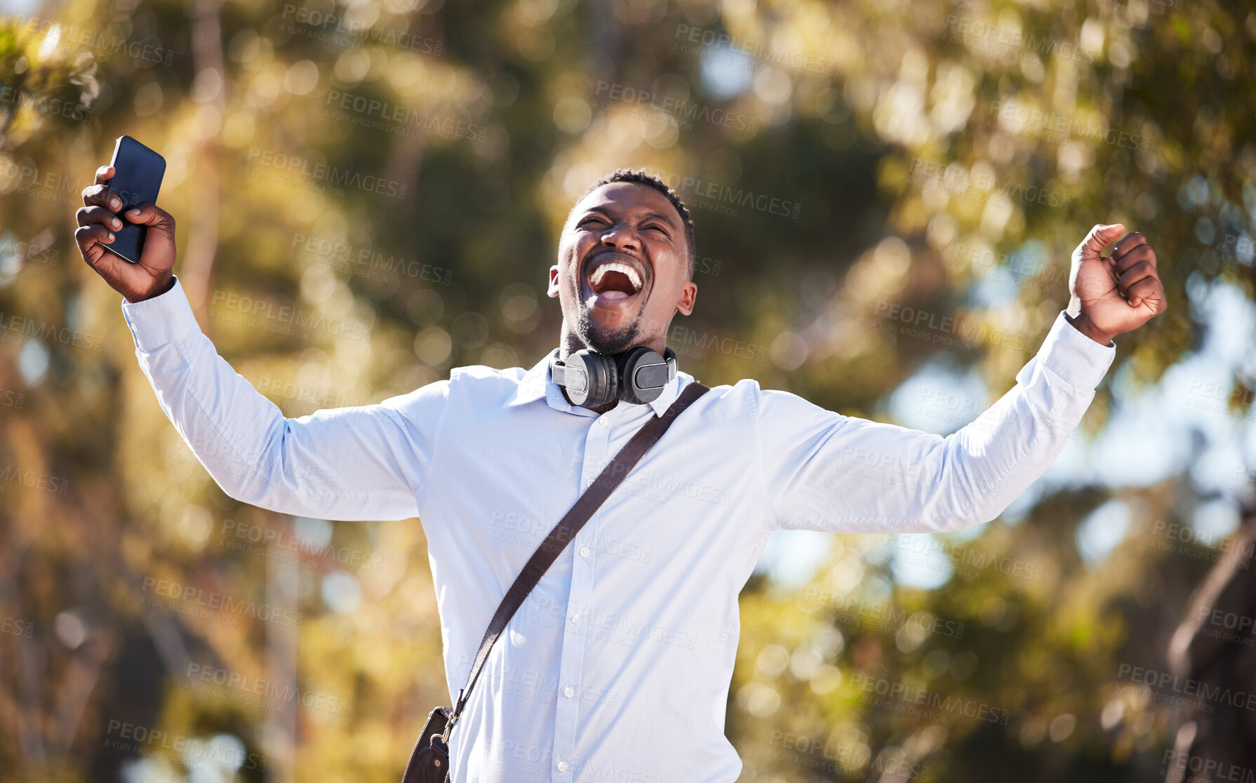 Buy stock photo African American man celebrating a win while using his cellphone and wearing headphones. Happy young male expressing success and victory while walking outside. Good news makes this man smile