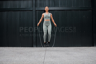 Buy stock photo One young african american female athlete skipping using jump rope while exercising outside in the city. Beautiful and dedicated mixed race sportswoman working out alone against an urban background