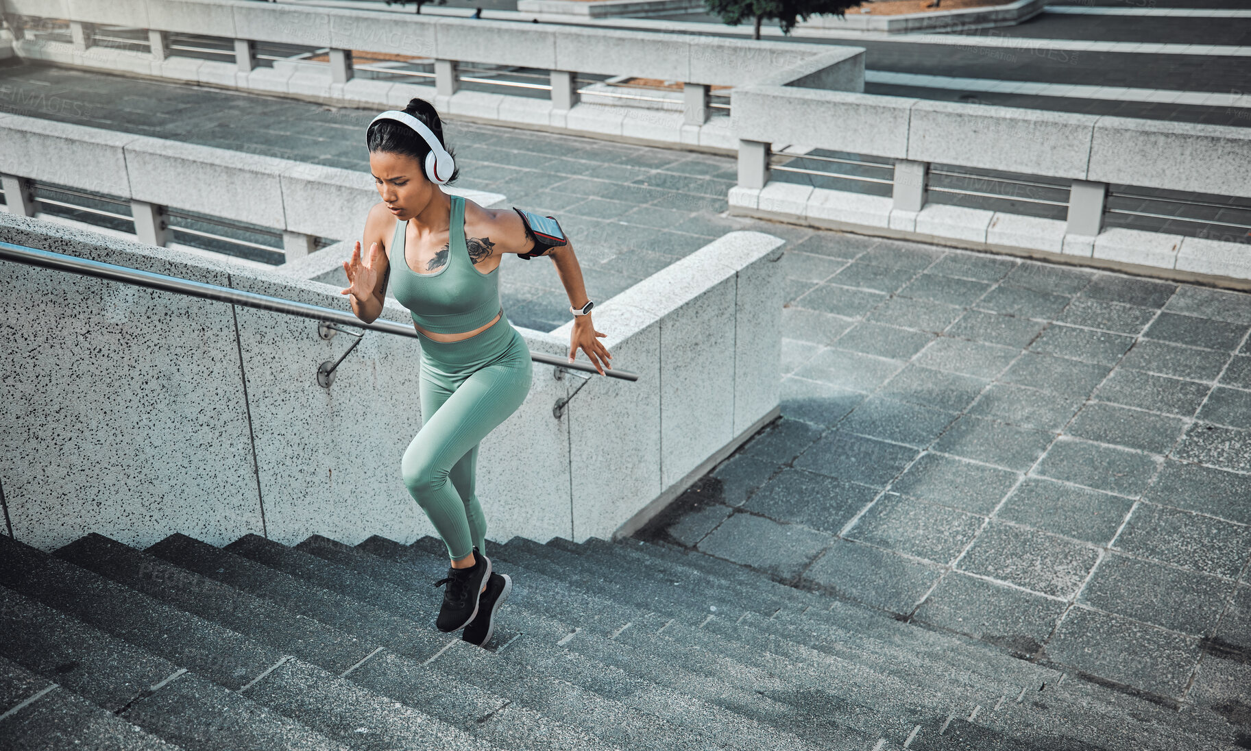 Buy stock photo Young mixed race female athlete wearing gymwear and headphones while running up the steps of a building outside. Young sportswoman focused on her speed, body, fitness and cardio health while training