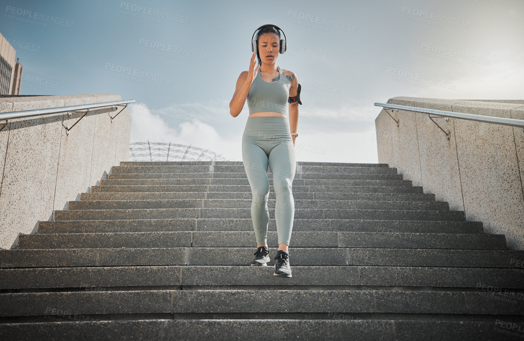 Buy stock photo Young mixed race female athlete listening to music on headphones while running down the steps of a building outside. Young female focused on her speed, body, fitness and cardio health while training