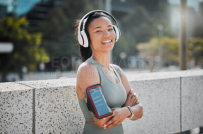 Buy stock photo Young mixed race female athlete wearing headphones armband while listening to music  and standing with her arms crossed exercising outside in the city. Positive, happy, workout