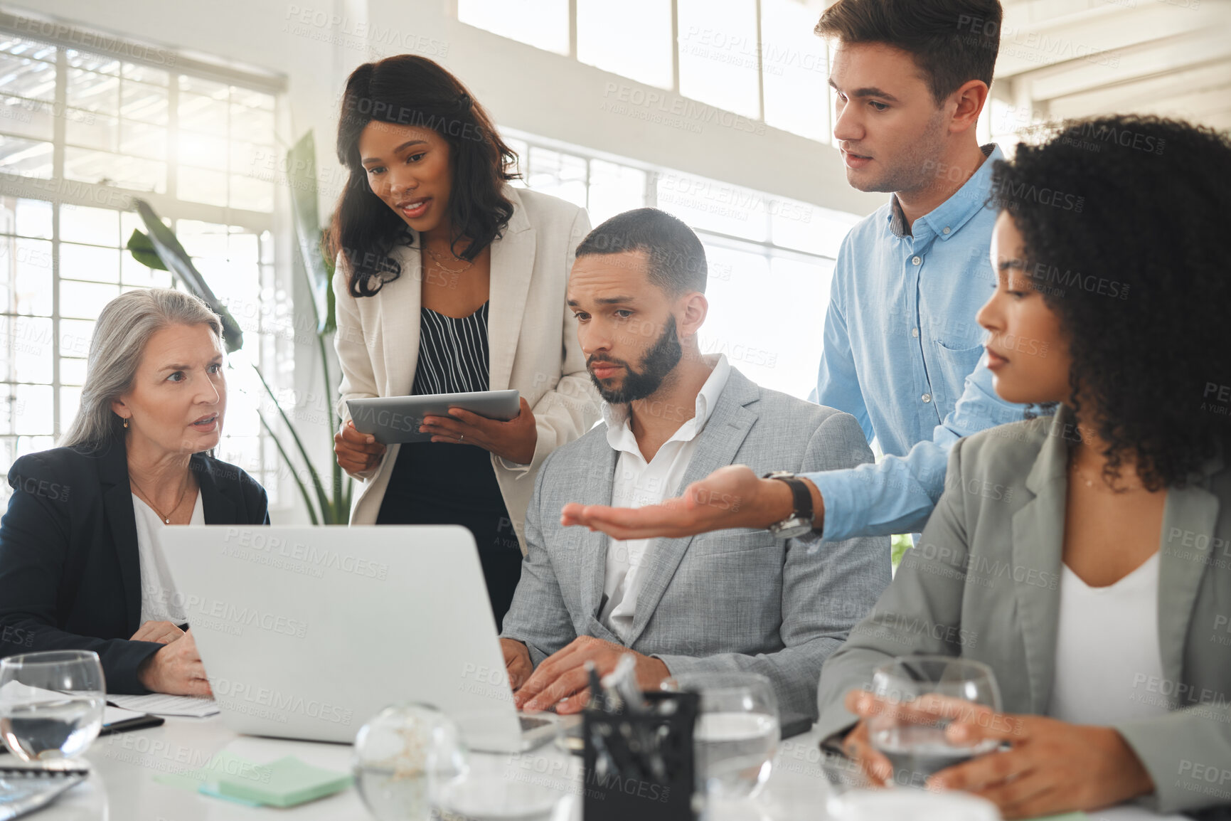 Buy stock photo Diverse group of businesspeople having a meeting while standing together at a table at work. Business professionals talking and planning while using a laptop in an office. Colleagues discussing a business strategy
