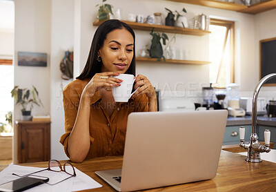 Young mixed race woman drinking a cup of coffee while using her laptop alone in the morning in the kitchen. Content hispanic female smiling and enjoying a cup of tea while reading an email on her computer at home