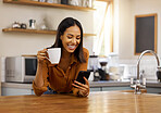 Young happy mixed race woman drinking a cup of coffee while using her phone alone in the morning in the kitchen. One cheerful hispanic female smiling and enjoying a cup of tea while using social media on her phone at home