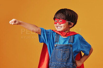 Adorable little asian boy wearing a mask and a cape while pretending to be a superhero against an orange studio background. Cute happy boy pretending to be a character for halloween