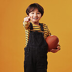 Cute little mixed race boy smiling and pointing while holding a football and posing against an orange copyspace background