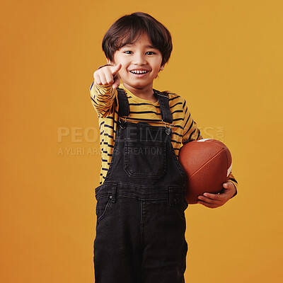Cute little mixed race boy smiling and pointing while holding a football and posing against an orange copyspace background