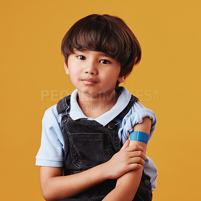 Portrait of a little asian boy holding his arm and showing his bandaid. Child wearing a plaster after being forced to get the vaccine, sad kid after getting an injection against an orange background