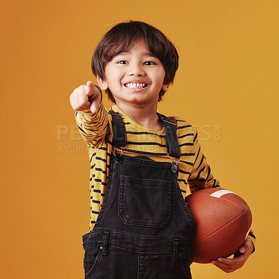 Cute little mixed race boy smiling and pointing while holding a football and posing against an orange copyspace background