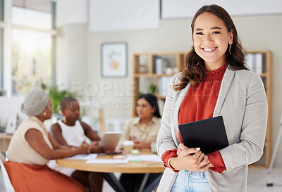 Smiling asian business woman holding a digital tablet while colleagues sit behind her in office. Ambitious and happy professional standing after browsing schedule on technology before leading meeting