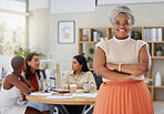 Portrait of smiling african american plus size business woman standing with her arms folded while colleagues sit behind her in office. Ambitious, confident, happy black professional with arms crossed